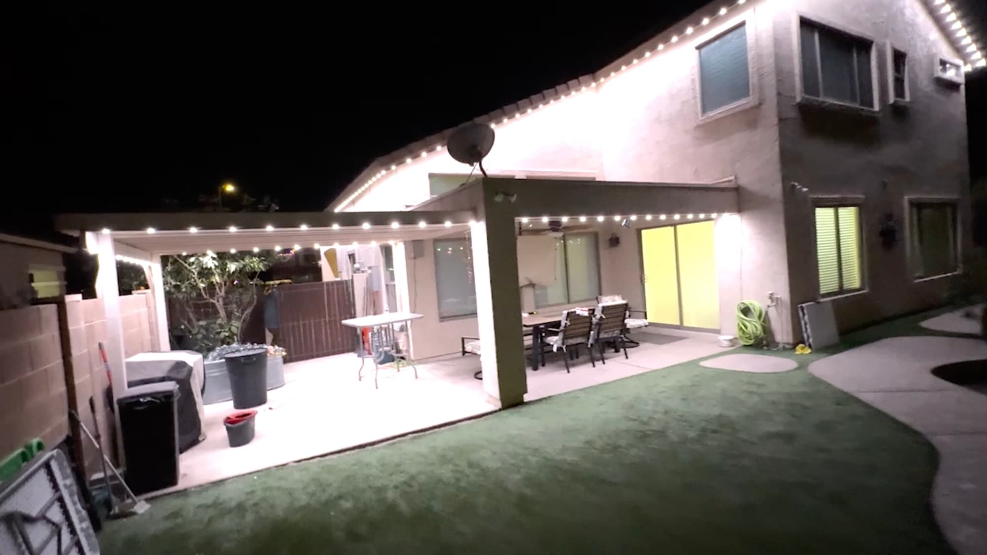 Backyard of a two-story stucco home at night, with white permanent lights along the edges of a patio cover and the upper roofline.