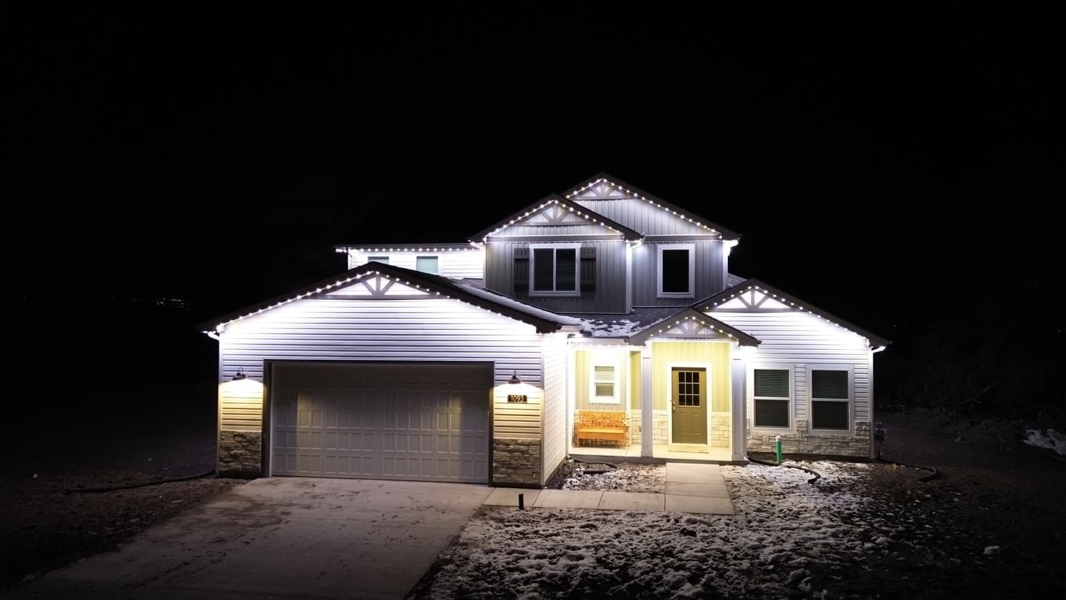 Two-story siding-and-stone home with bright white permanent lights outlining nested gables on a winter night.