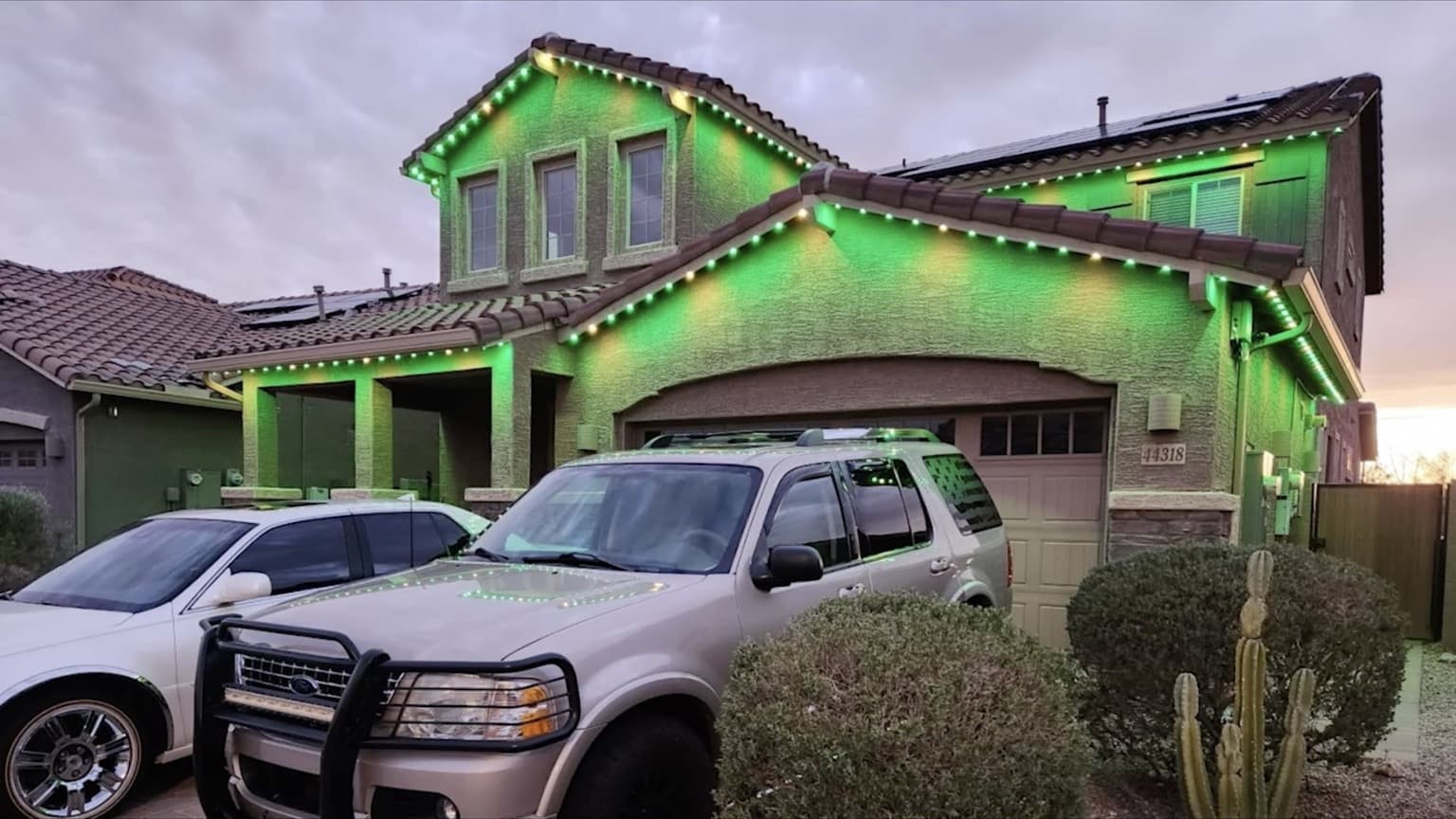 Two-story stucco home with tile roof, solar panels, and green-and-white permanent lighting along layered eaves and gables.